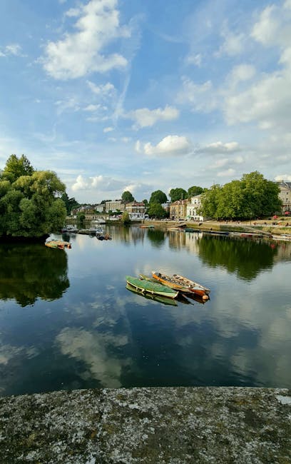 A scenic view of a calm river during daytime, reflecting the partly cloudy sky and surrounding greenery. Several small boats, made of wood and plastic, are moored along the riverbank, with some tied to each other. On the opposite side of the river, a row of residential buildings and houses with light-colored facades and pitched roofs are visible, interspersed with trees and landscaped areas. The scene suggests a peaceful setting suitable for home relocation or moving services, with the river and boats emphasizing transportation and logistics in the context of house removals. The lighting indicates a bright, clear day, and the environment appears quiet and suburban. This image could be used on the Kingston house removals page of removalskingston.co.uk to illustrate the scenic locale near Kingston Bridge and the logistical environment for furniture transport and packing during a move.