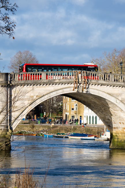 A red and white bus travelling across a stone arch bridge over a river in Kingston, with pedestrians walking along the riverside path and buildings visible in the background. The bridge's decorative railing is made of stone and concrete, and the river below reflects the sky and boats moored along the bank. The surrounding trees are mostly leafless, indicating a late autumn or winter season. This scene illustrates an urban environment commonly associated with house removals and furniture transport, with the bridge serving as a key landmark in Kingston near Kingston Bridge. The image captures a daytime setting with clear weather, suitable for moving or relocation services by companies like Removals Kingston, which specialise in house removals near the area.